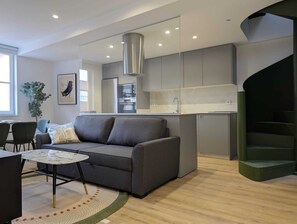 Una sala moderna con un sofá gris, una mesa de centro redonda y una pequeña mesa de comedor. Se ve una cocina con gabinetes grises y electrodomésticos de acero inoxidable al fondo.