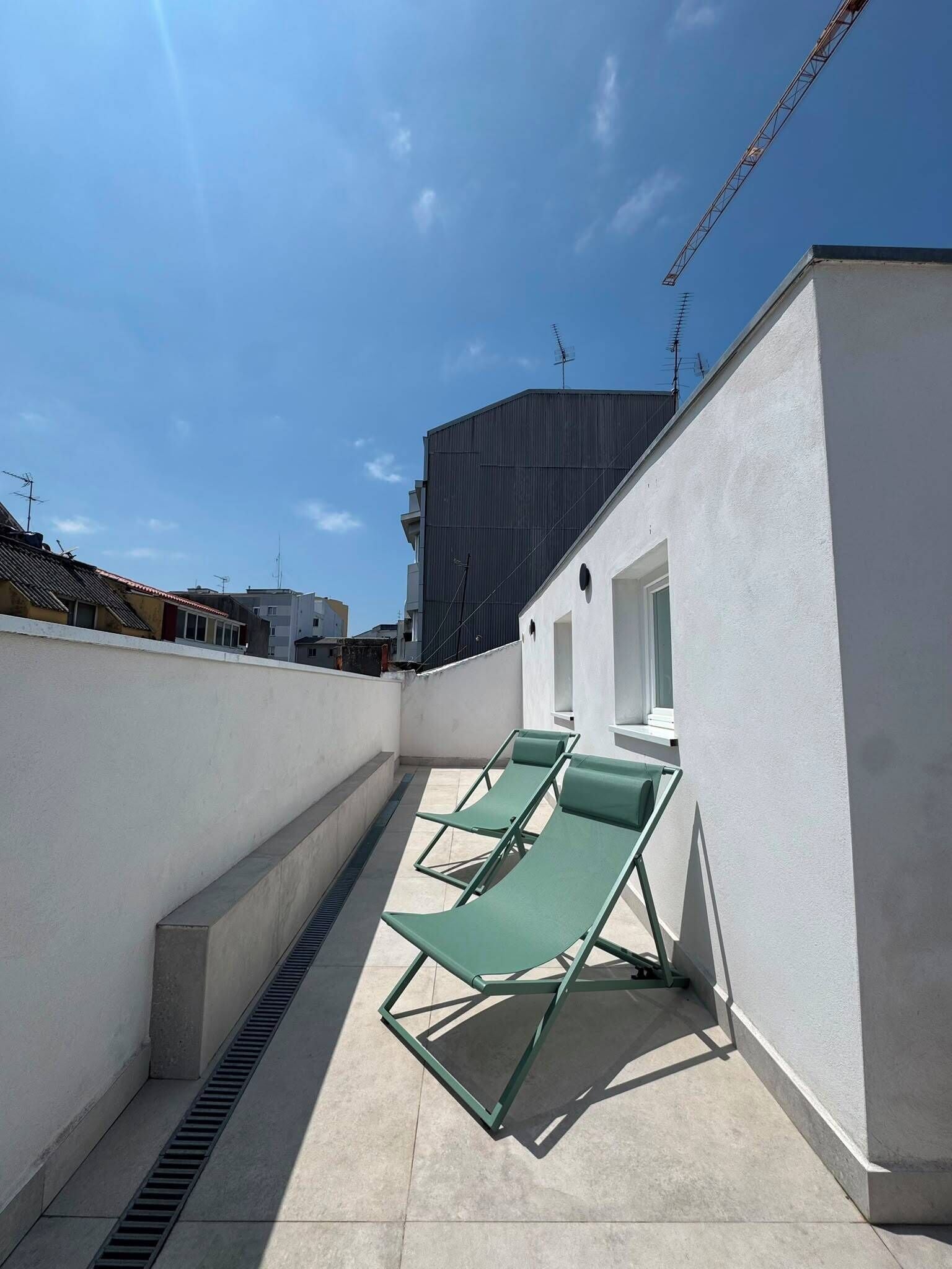A rooftop terrace with two green lounge chairs, a white building, and a clear blue sky.