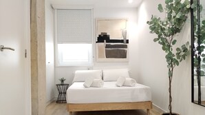 A bedroom with a bed, a window with blinds, a small table with a plant, and a potted tree. - Paz Apartments By Como En Casa (A CORUÑA)