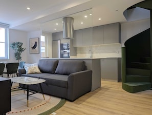 A modern living room with a grey sofa, a round coffee table, and a small dining table. A kitchen with grey cabinets and stainless steel appliances is visible in the background. - Paz Apartments By Como En Casa (A CORUÑA)
