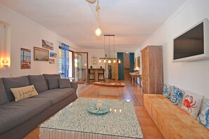A living room with a sofa, a coffee table, and a TV. There are pictures on the wall and a dining area in the background.