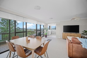A modern living room with a large TV, a dining area with chairs, and a balcony with a view. - Coastal Apartment with Ocean Views, Pool, Sauna & Tennis Court (Forster)