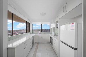 A modern kitchen with white cabinetry, a built-in oven, a refrigerator, and a window with a city view. - Coastal Apartment with Ocean Views, Pool, Sauna & Tennis Court (Forster)