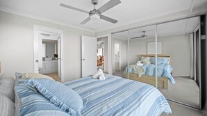 Une chambre à coucher comprenant un lit, un ventilateur de plafond, un grand miroir et une porte coulissante donnant sur une autre pièce.