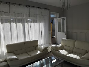 Un salon avec deux canapĂ©s en cuir beige, une table basse en verre, des rideaux blancs et transparents, et une porte donnant sur lâextĂ©rieur.