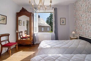 A bedroom with a large bed, a wooden chair, a mirror, and a view of a house through a window. - Holiday Home “Les Glycines” with Private Terrace and Garden (Rouffignac-Saint-Cernin-de-Reilhac)