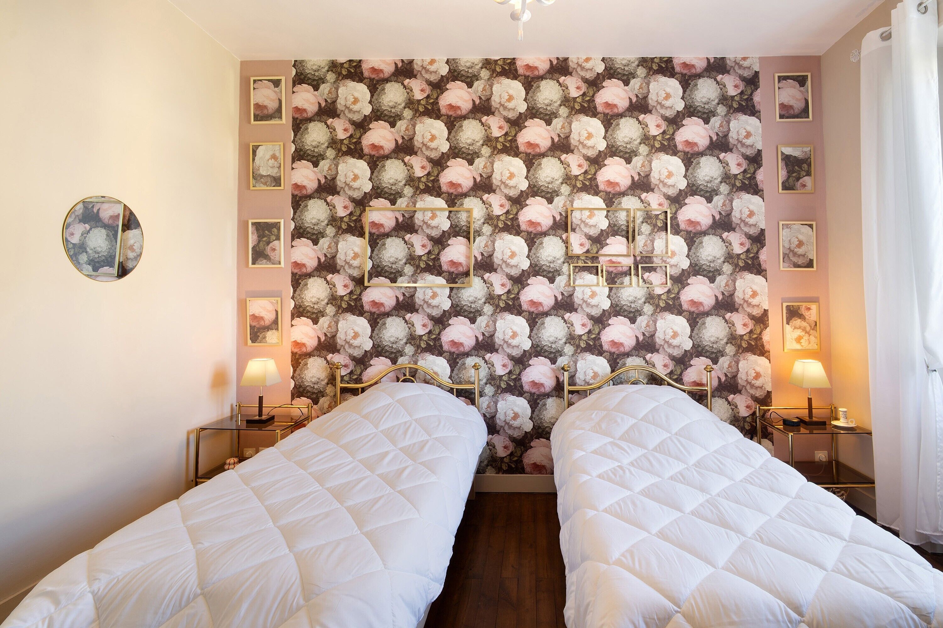 Ein Schlafzimmer mit zwei Betten, floralem Wandpapier, Nachttischlampen und einem runden Spiegel.