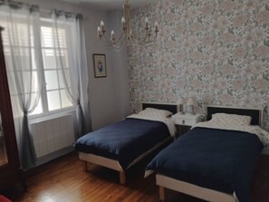 A bedroom with two beds, a chandelier, a window with curtains, and floral wallpaper.