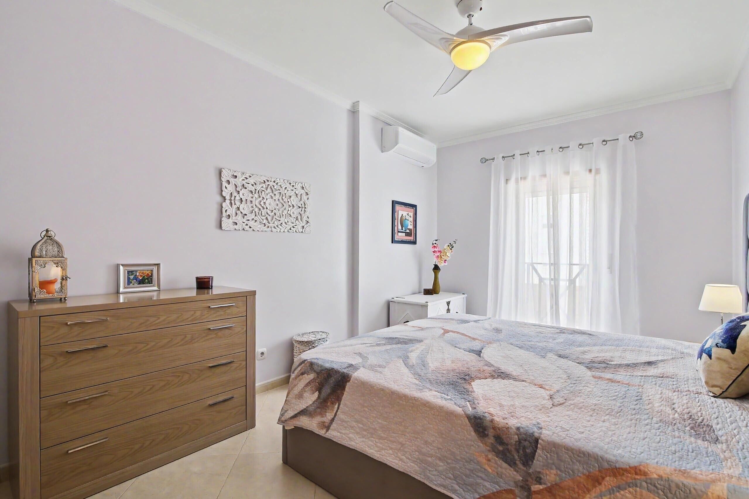 A bedroom with a bed, a wooden dresser, a ceiling fan, a window with curtains, and a bedside table with a lamp.