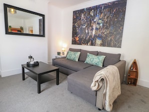 A living room with a grey sofa, a coffee table, a mirror, a night cityscape painting, a lamp, and a small wooden shelf.