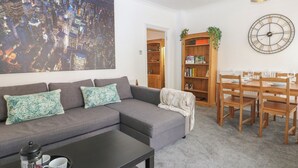 A living room with a gray sofa, a coffee table, a cityscape painting, a wooden bookshelf, a dining table with chairs, and a large wall clock. - Foreshore (Deal)