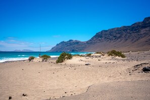 A beach with a clear sky, rocky terrain, and a flagpole. - Quicksand • Ocean views • Beach 1 min • LVH (Famara)