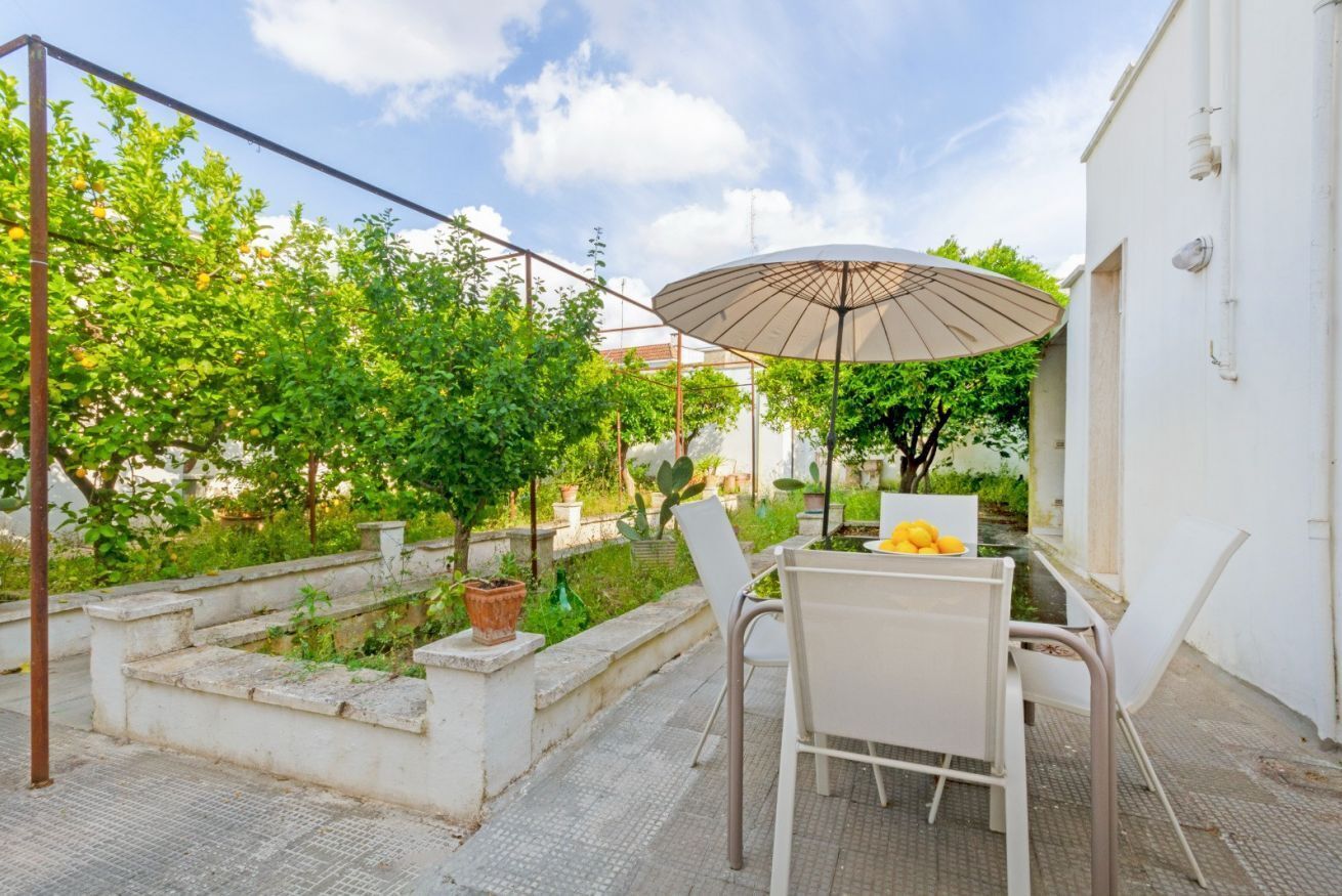 Une terrasse avec une table et des chaises, un grand parasol et un jardin agrémenté d’arbres et de plantes.