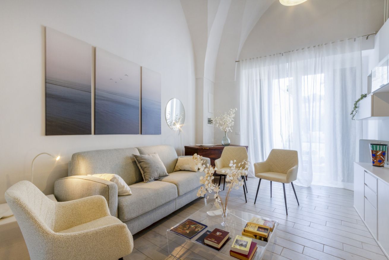 A modern living room with a sofa, armchairs, a coffee table with books, a mirror, and artwork on the wall.