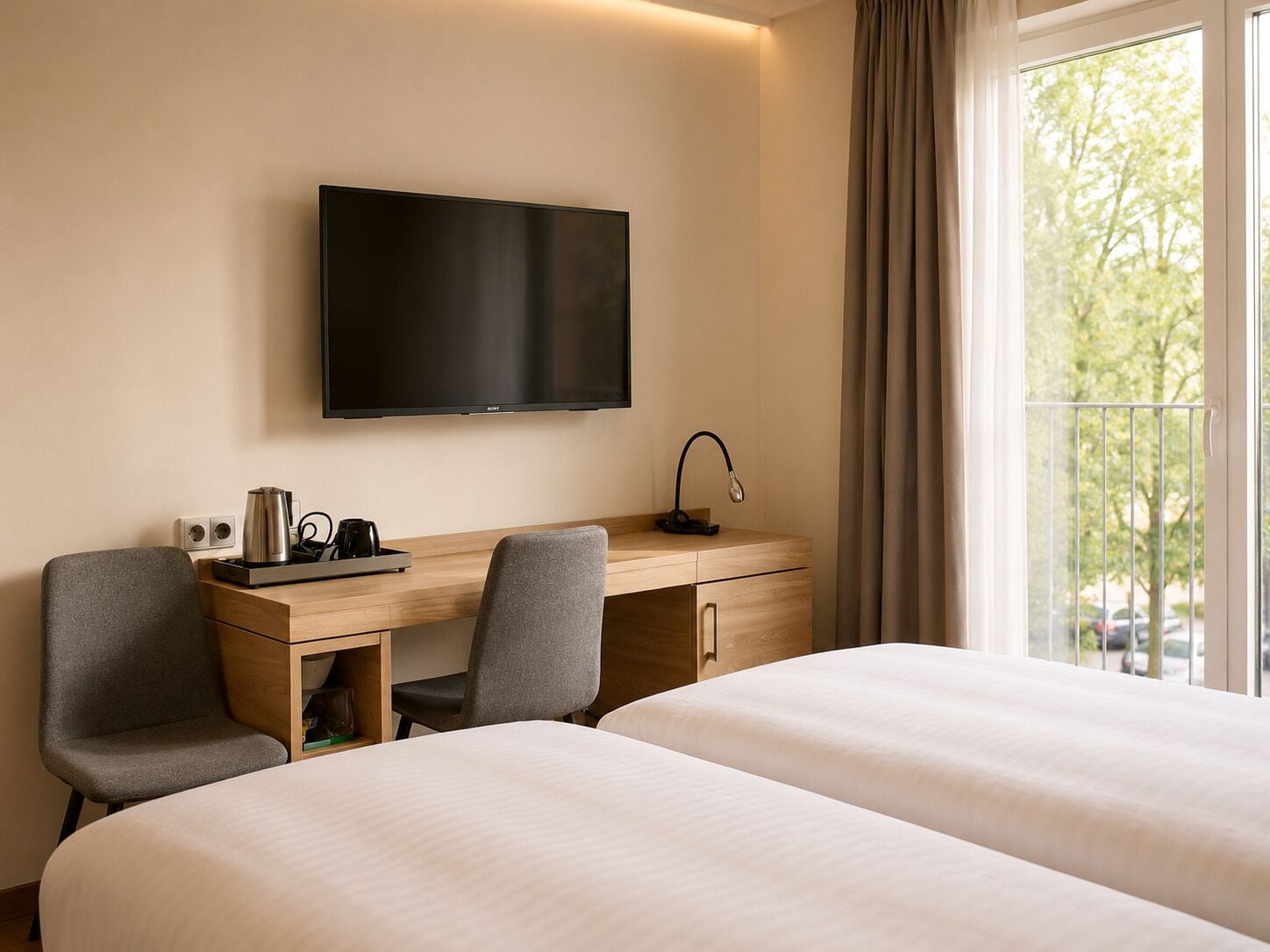 A hotel room with two beds, a desk with a TV, a lamp, and a window with a view of trees.