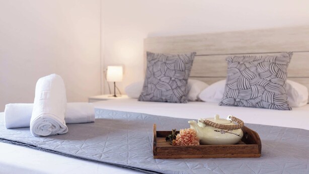 Ein ordentlich bezogenes Bett mit aufgerollten weißen Handtüchern, ein Holztablett mit Teekanne und Blumen sowie gemusterte Kissen.