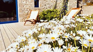 Un bâtiment en pierre doté d’une terrasse en bois, de deux chaises longues et d’un jardin rempli de fleurs blanches.