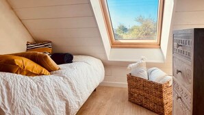 Une chambre avec un plafond en pente, un puits de lumière, un lit avec une literie blanche, un panier en osier et une commode en bois.