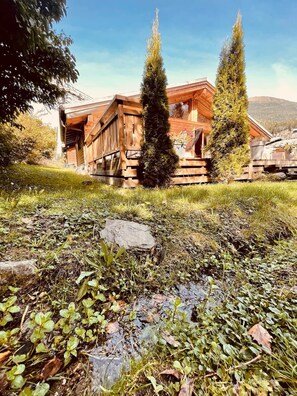 A rustic wooden cabin with a sloped roof, surrounded by a grassy area with rocks and plants.