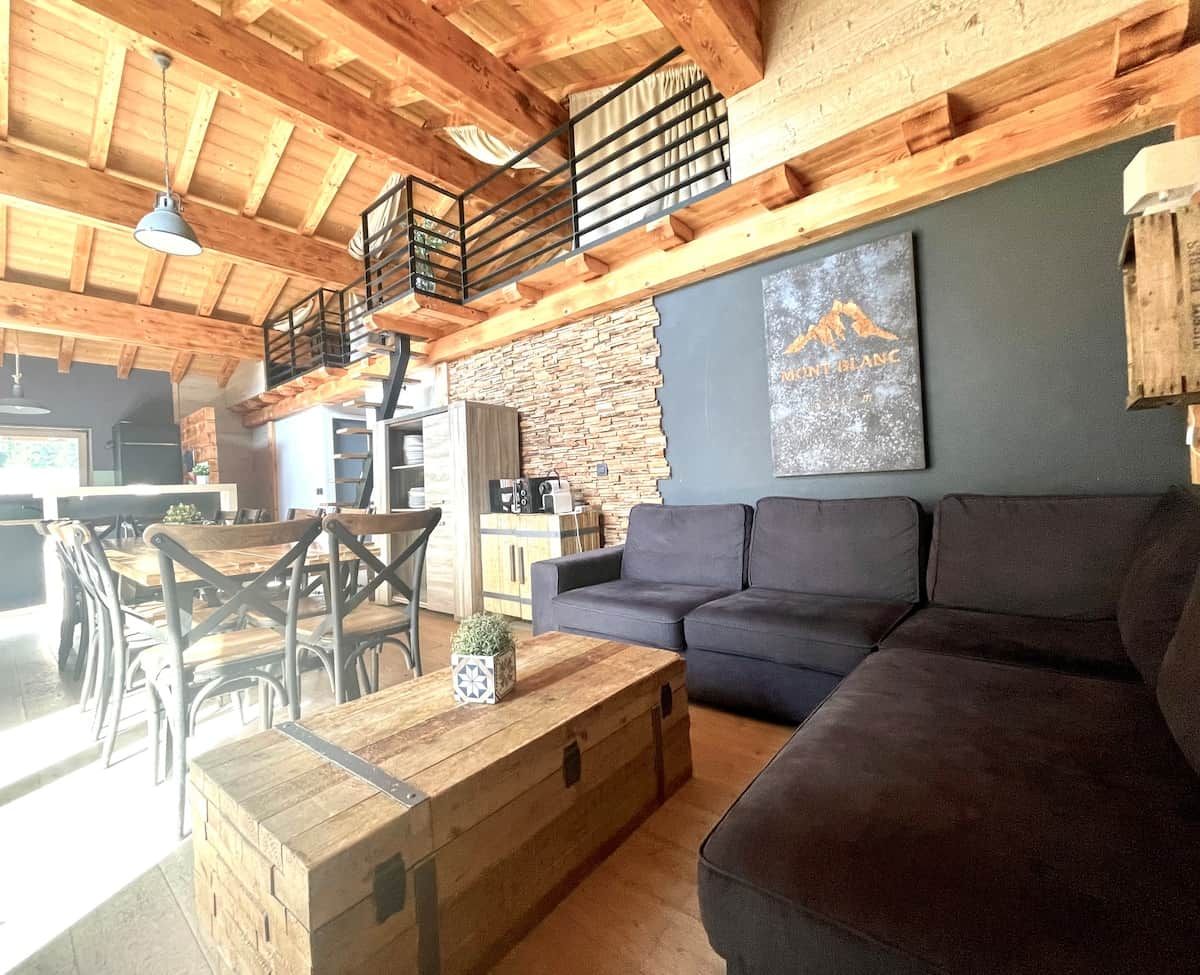 Un interior rústico con techo de madera, pared de piedra, sofá gris oscuro y una zona de comedor con mesas y sillas de madera.