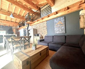 A rustic interior with a wooden ceiling, a stone wall, a dark gray sofa, and a dining area with wooden tables and chairs.