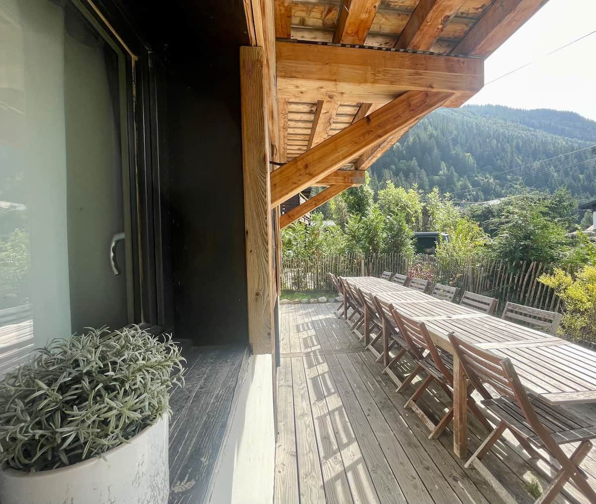 Terraza de madera con mesa y sillas, una planta en maceta y vistas a una montaña boscosa.