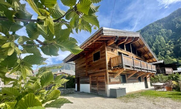 A wooden chalet with a balcony, surrounded by greenery and mountains.