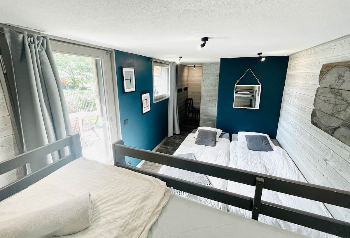 Ein Zimmer mit zwei Betten, einem fensterbelaubten Fenster, einem kleinen Tisch und einem an der Wand befestigten Regal.
