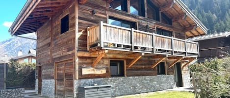 A wooden chalet with a balcony, stone foundation, and a mountainous backdrop.