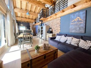 A spacious living area with a wooden ceiling, a dark sofa, a dining table, and a kitchen in the background. - Chalet 14 pers, jacuzzi, MontBlanc view, 5 bedrooms (Chamonix)