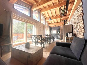 A spacious living area with a wooden ceiling, large windows, a stone wall, and a dining area with chairs and tables.