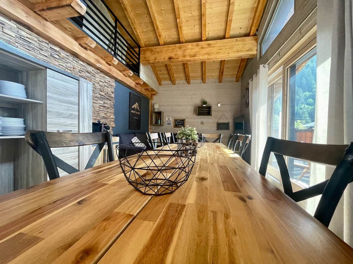 Una mesa de comedor de madera con un centro geométrico, rodeada de sillas, en una habitación con vigas de madera a la vista y ventanas grandes.