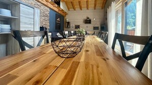 Una mesa de comedor de madera con un centro geométrico, rodeada de sillas, en una habitación con vigas de madera a la vista y ventanas grandes.