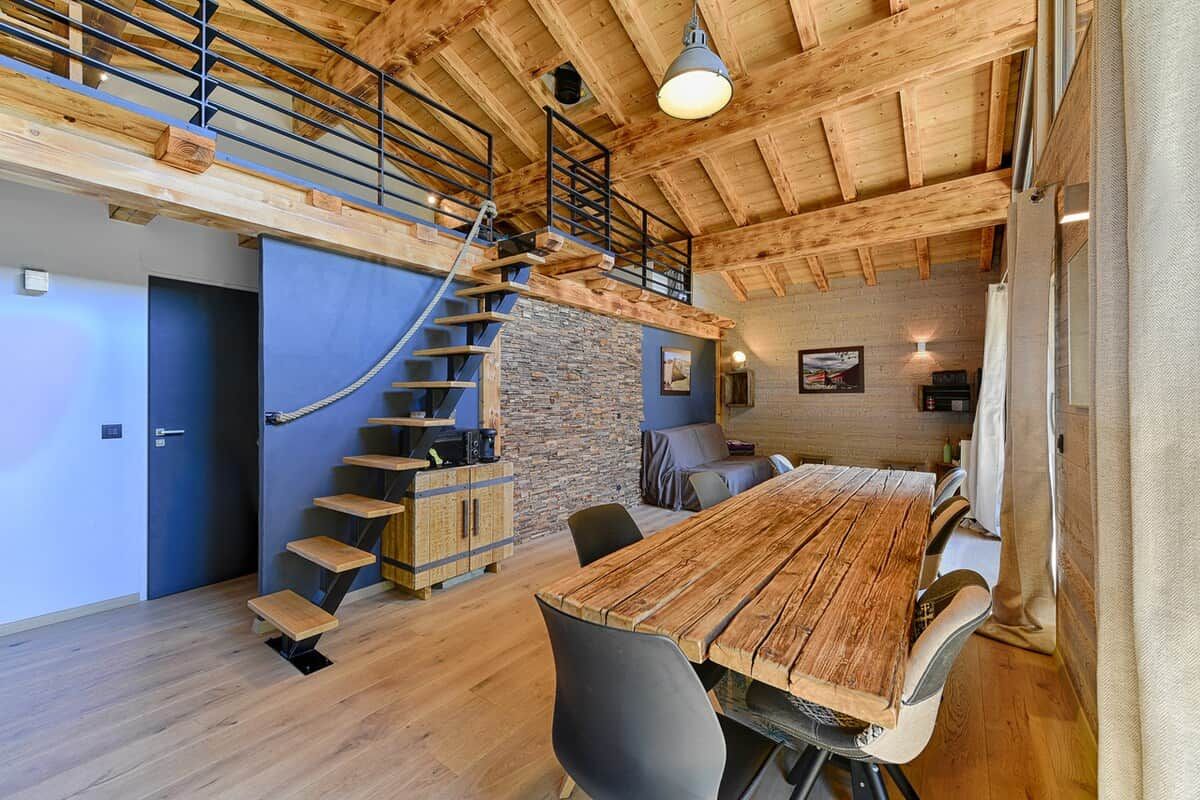 Una habitación amplia con una escalera de madera, una larga mesa de comedor y una pared de piedra.