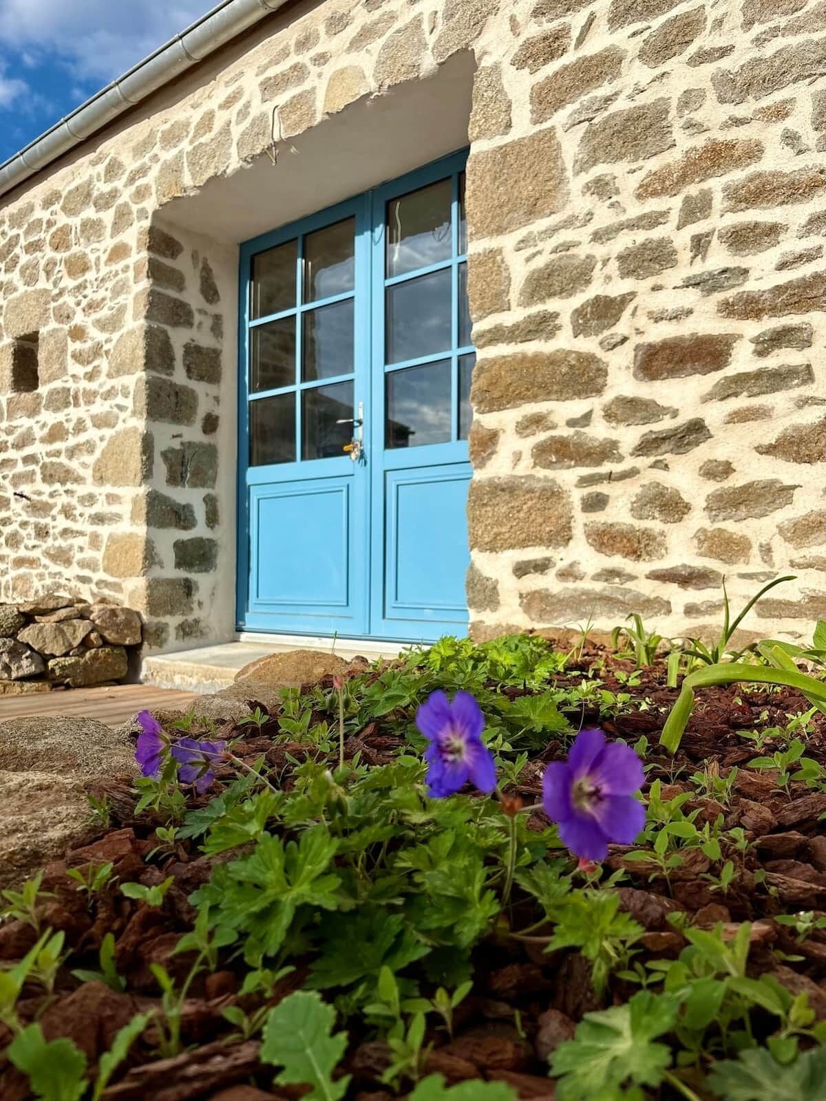 Un bâtiment en pierre avec une porte bleue et des volets aux fenêtres, entouré d’un jardin où poussent des fleurs violettes.