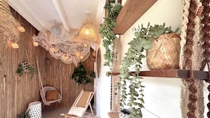 Um quarto aconchegante com teto de madeira, uma poltrona estofada, uma mesinha e uma prateleira com plantas.