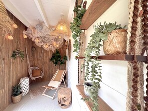 A cozy room with a wooden ceiling, a cushioned chair, a small table, and a shelf with plants.