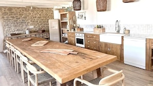 Uma cozinha com mesa de madeira, cadeiras, parede de pedra, armários de madeira, geladeira branca e cestos de vime pendurados.