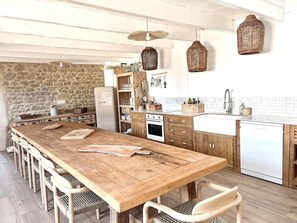 A kitchen with a wooden table, chairs, a stone wall, wooden cabinets, a white refrigerator, and hanging wicker baskets.