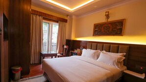 A hotel room with a large bed, a nightstand, a desk with a chair, a window with curtains, and a wall-mounted artwork. - Boudha Boutique Inn (Kathmandu)