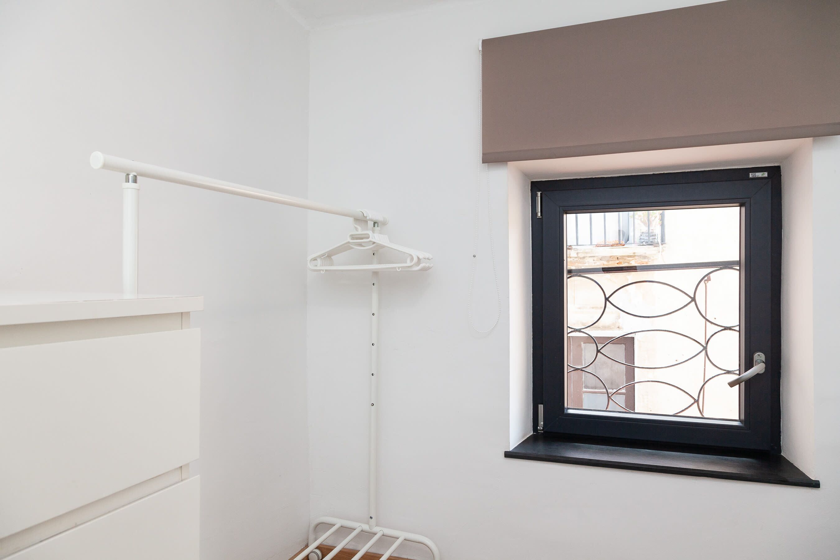 A room with a white wardrobe, a hanging rack, a window with a metal grill, and a door.