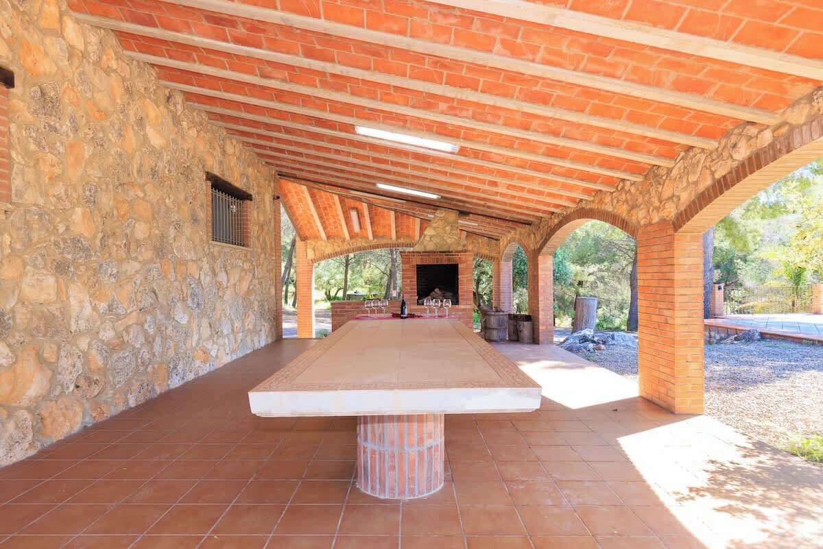 A covered outdoor dining area with a stone wall, a long table, and a brick ceiling.