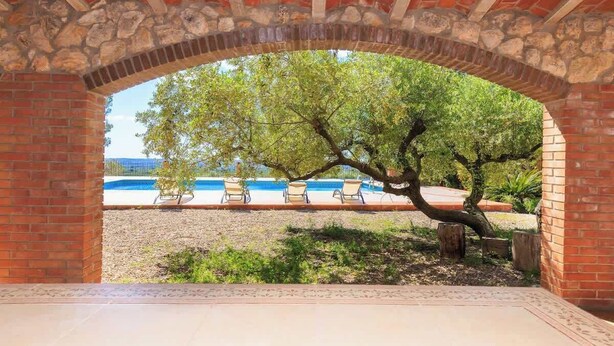 An outdoor patio area with a brick archway, a tree, and a pool with lounge chairs. - Masia Garriga (Tarragona)