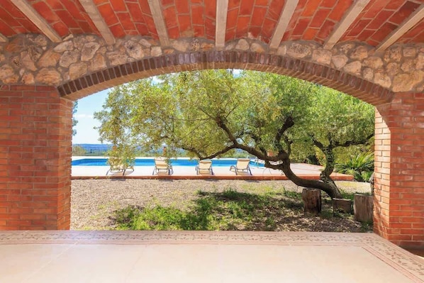 Un espace extérieur aménagé en terrasse, avec une arche en briques, un arbre et une piscine entourée de chaises longues.