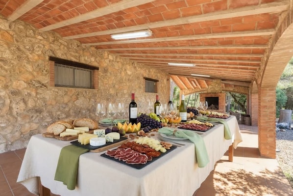 A covered outdoor dining area with stone walls, a long table set with various cheeses, meats, fruits, and wine glasses.