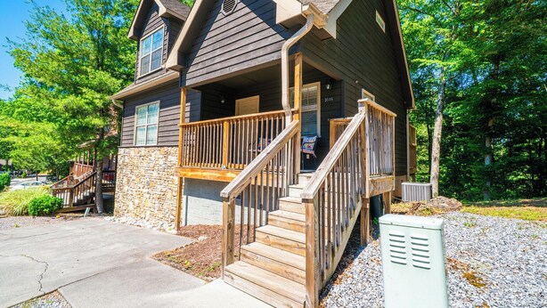 A two-story house with a stone foundation, a wooden porch with a railing, and a driveway leading to the entrance. - Bear Cozy: Snuggle Up in the Smokies (Pigeon Forge)