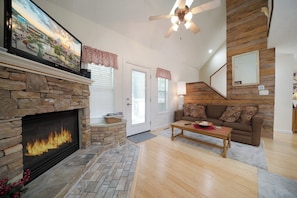 A living room with a stone fireplace, a ceiling fan, a couch, and a television. - Beary Cozy | Hot Tub | Both Beds w/ Private Bath | Sleeps 6 (Pigeon Forge)