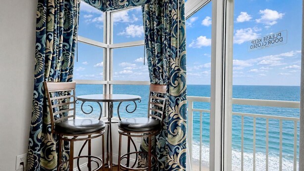 Ein Eckbalkon mit Meerblick, zwei Stühle, ein kleiner Tisch und bunte Vorhänge.