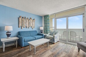 A room with a blue wall, a blue sofa, a white table, a patterned armchair, and a balcony with white chairs. - Oceanfront 2BR - 19th Floor w/ King & 2 Doubles + Breakfast for 2 + Hut Tub (Myrtle Beach)