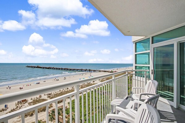 Ein Balkon mit weißen Stühlen, Blick auf den Strand und einen sich ins Meer erstreckenden Steg.